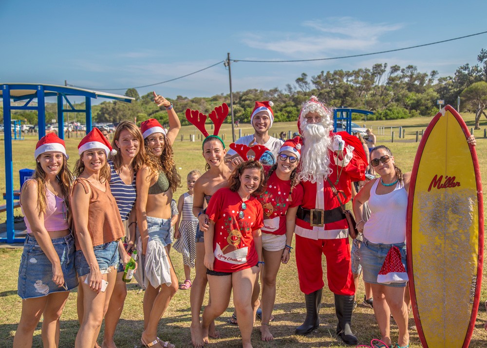 grupo de estudiantes sonrientes en una Barbacoa con gorritos de navidad y el Papa Noel. Una estudiante sujeta una tabla de surf