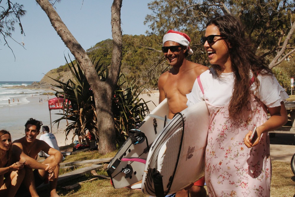 estudiantes sonrientes con una tabla de surf en una playa de Sydney y con el gorrito de Navidad