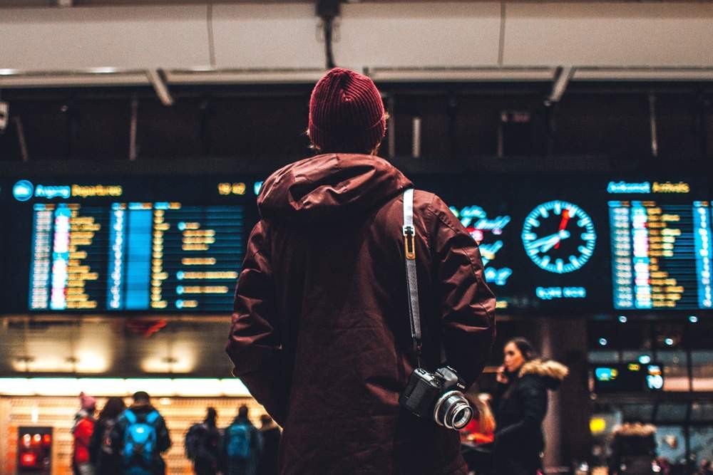 chico con gorro de lana granate y chaqueta de espaldas mirando el panel del aeropuerto. Lleva una cámara de fotos colgada en el hombro