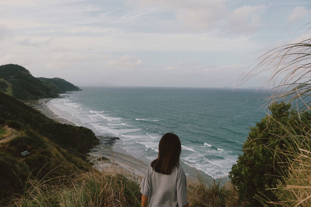 chica morena de espaldas mirando al mar y rodeada de vegetación