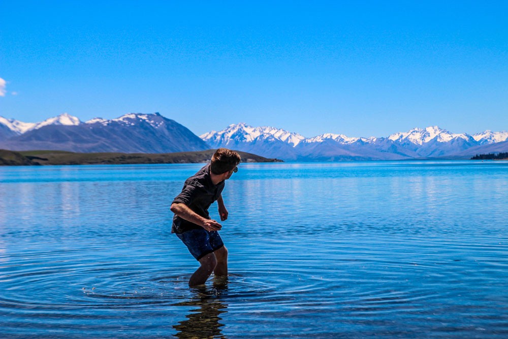 chico de espaldas tirando una piedra en un lago y detrás montañas