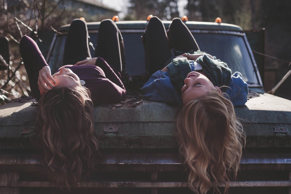 dos chicas tumbadas de boca para abajo encima de un coche antiguo. Se miran entre ellas riendo