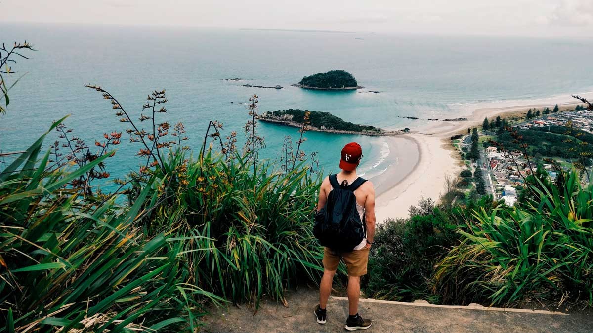 chico de espaldas y en una montaña observando el mar de Nueva Zelanda. Lleva una mochila negra y una gorra de color rojo