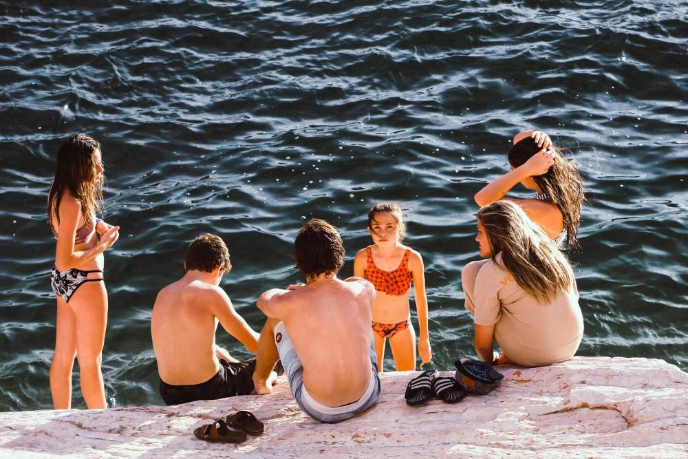 Grupo de adolescente en el que hay 4 chicas y 2 chicos en traje de baño y están sentados en una roca delante del mar. 