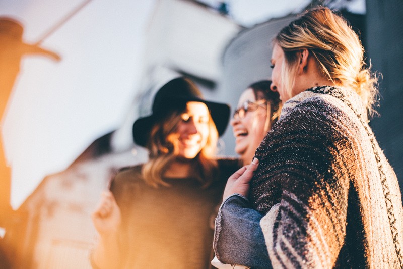 tres chicas mirándose entre ellas y riendo. Detrás de ellas hay edificios