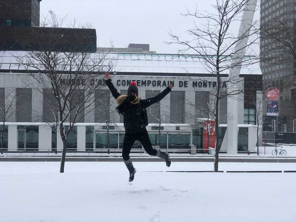 chica rubia abrigada saltando y detrás edificios y paisaje nevado