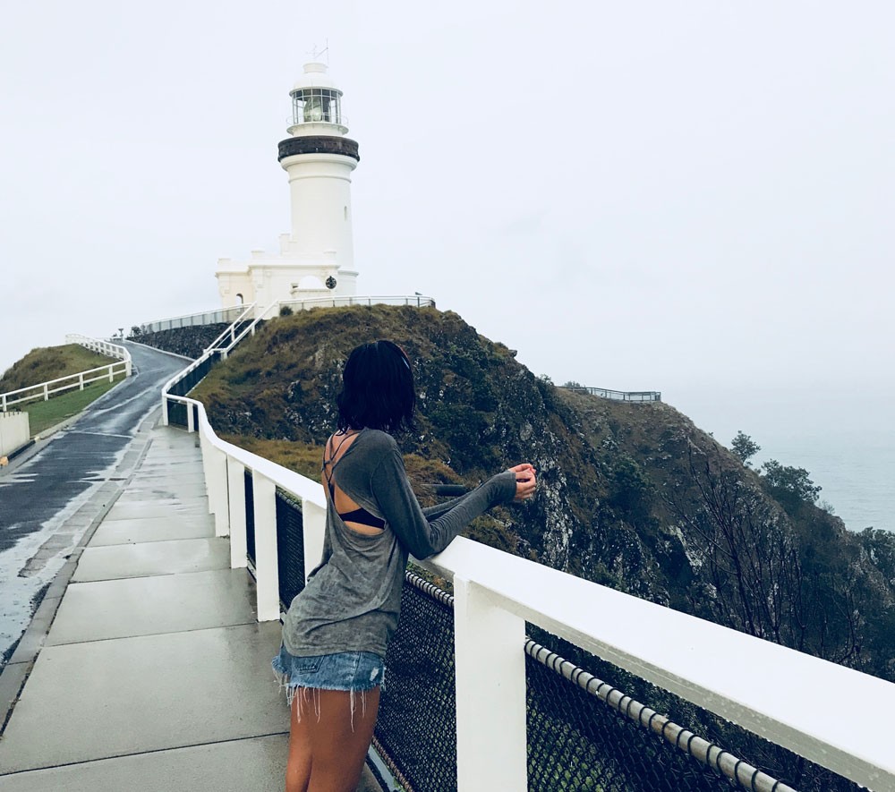 chica morena de espaldas en un puente que lleva a un faro 