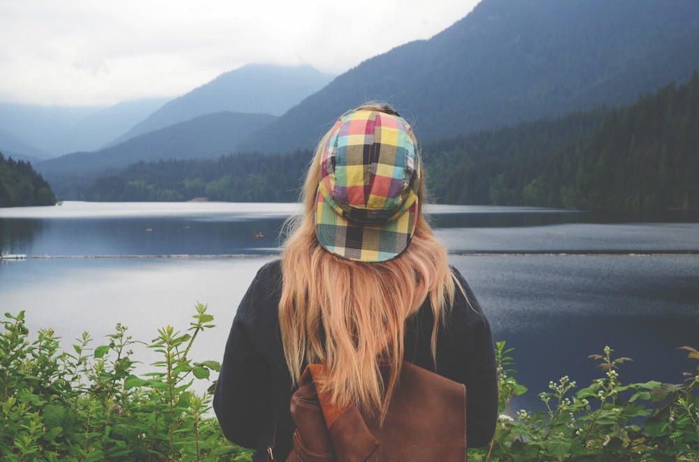 chica rubia de espaldas con una gorra de cuadros de colores y una mochila de piel marrón mirando un lago de Canadá