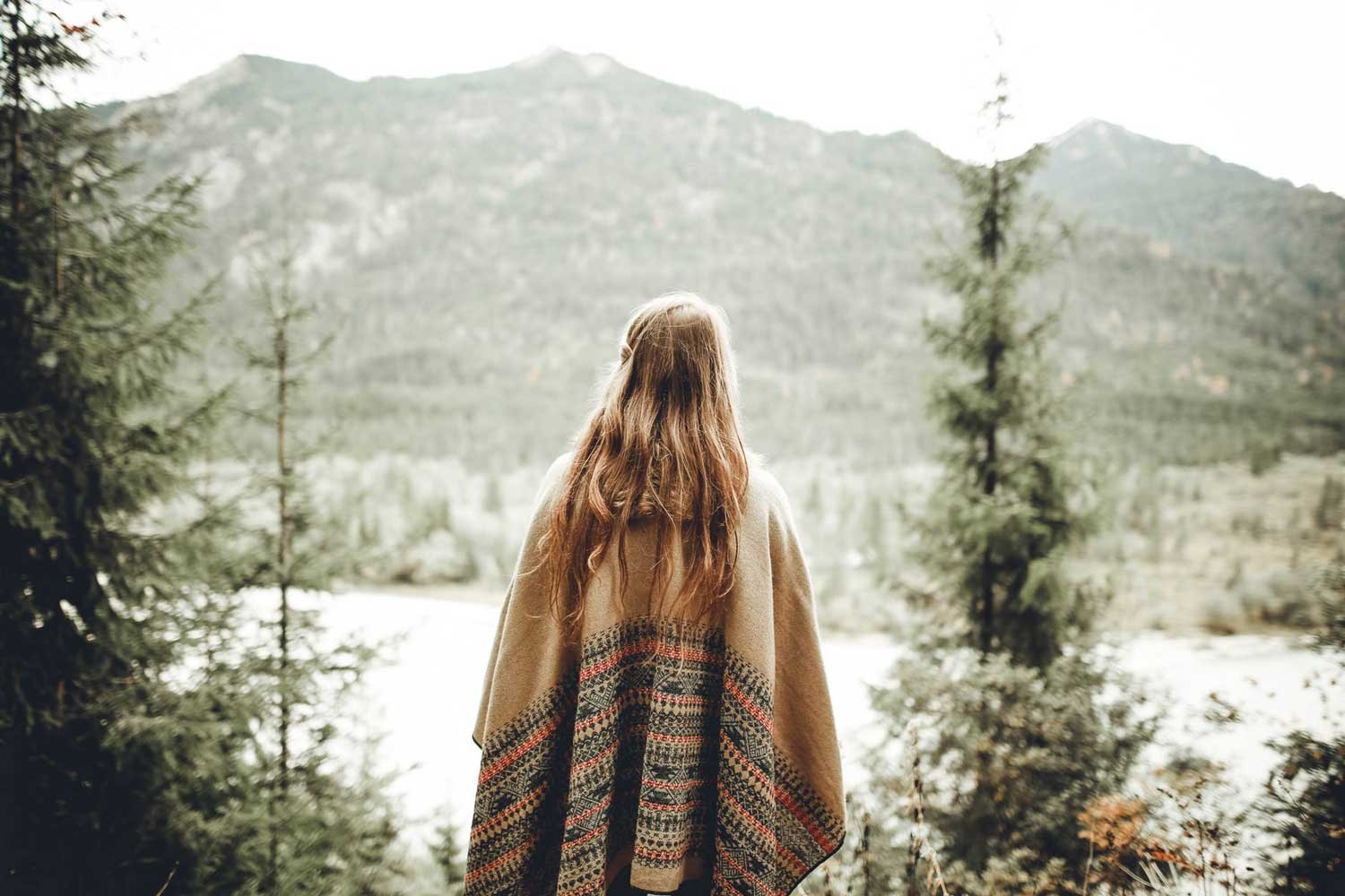 chica rubia con un poncho de color marrón de espaldas y enfrente suyo la naturaleza