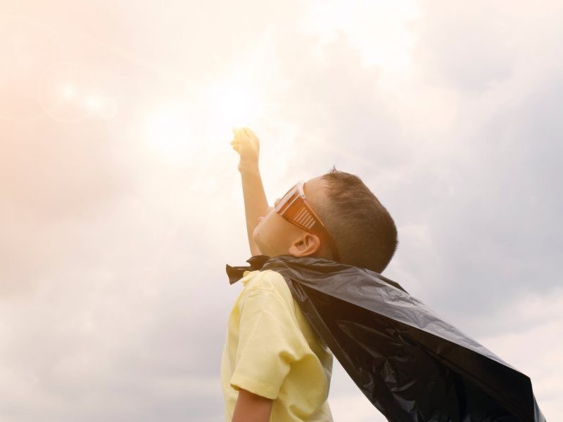 Niño con gafas rojas de super héroe con una capa negra y mirando al cielo con el brazo levantado 