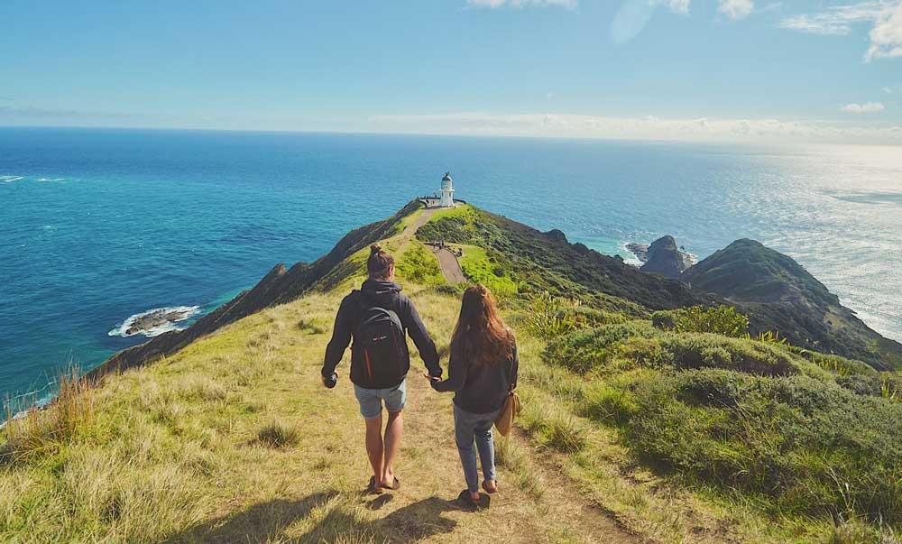 pareja de espaldas andando por una montaña y al final un faro