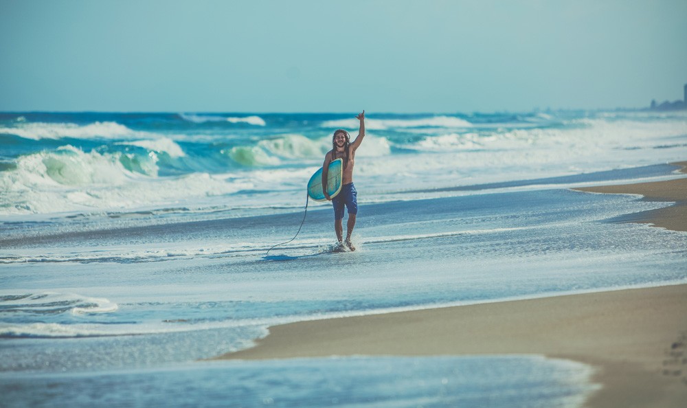 chico con el traje de baño y una tabla de surf saliendo del agua y sonriendo a cámara