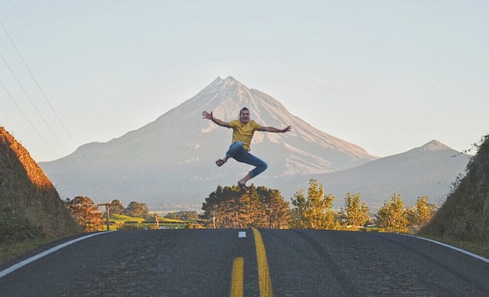 chico moreno saltando en la carretera y detrás una montaña