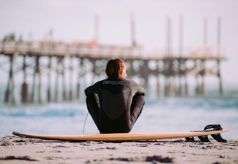 chico de espaldas con el neopreno y sentado encima de una tabla de surf