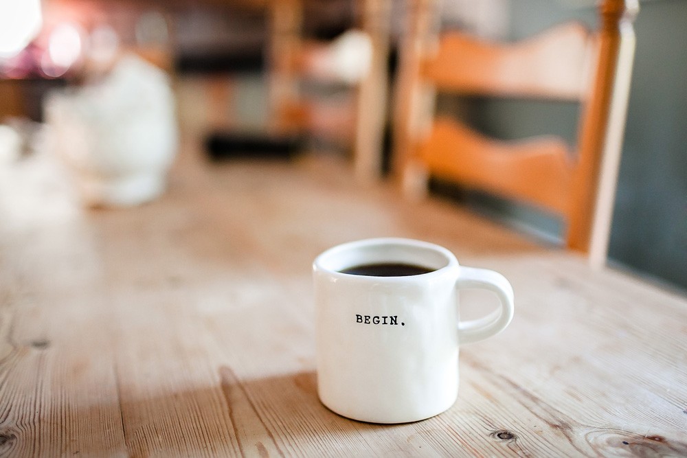 fotografía de una taza blanca con café encima de una mesa de madera. En la taza pone "begin"