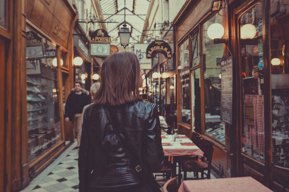 chica morena de espaldas con una chaqueta negra andando por un centro comercial y mirando el escaparate de un restaurante