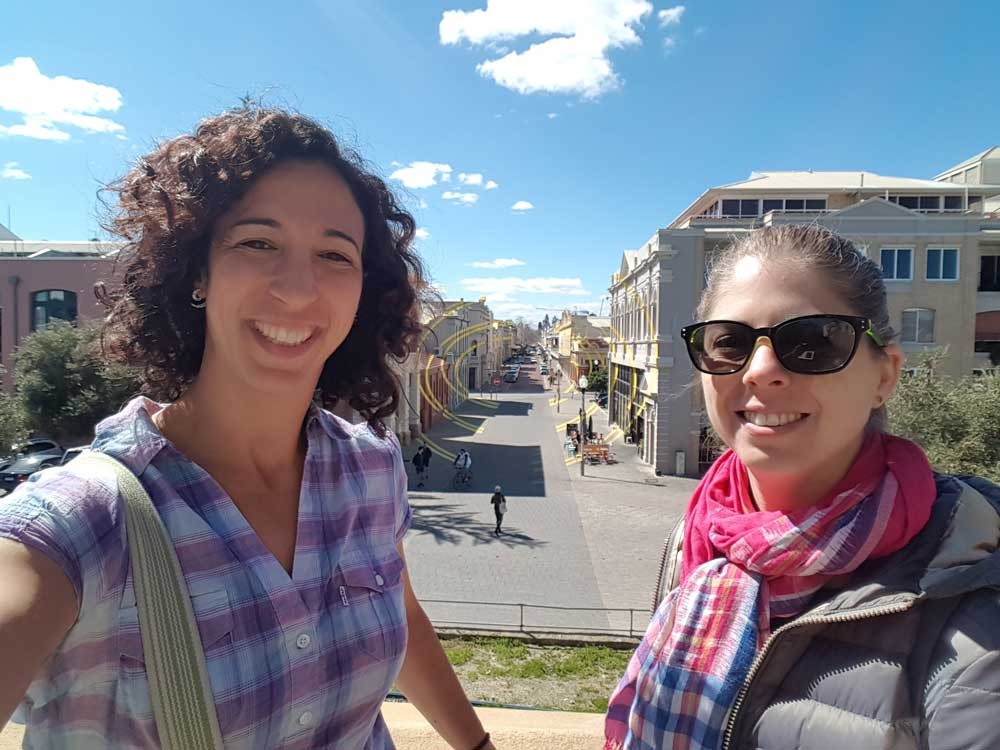 dos chicas morenas sonrientes mirando a cámara y detrás edificios